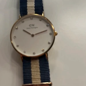 Klocka från Daniel Wellington - Säljer en stilren klocka från Daniel Wellington med en vit urtavla och guldfärgad boett. Klockan har en blå och vit randig textilrem som ger en klassisk look. Perfekt accessoar för att lyfta din stil. Obs batteriet i själva uret behövs uppdateras