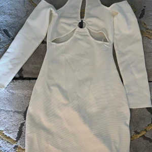 Vit klänning - Elegant vit klänning som är långärmad. Klänningen har en unik detalj framtill med en ring och utskärningar som ger en modern touch. Perfekt för speciella tillfällen.