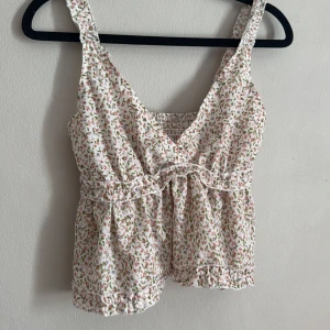 Blommig ärmlös topp - Söt blommig ärmlös topp med v-ringning och volangdetaljer. Perfekt för varma sommardagar. Den har en elastisk midja för en bekväm passform.