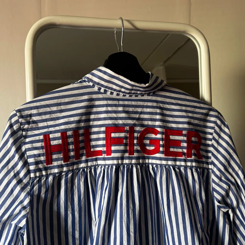 Säljer en stilren blå och vit randig skjorta från Tommy Hilfiger. Skjortan har en klassisk krage och knappar framtill. På baksidan finns en stor röd Hilfiger-logga. Perfekt för en avslappnad men ändå snygg look.. Paidat.