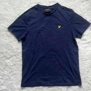 Mörkblå t-shirt från Lyle & Scott - Säljer en stilren mörkblå t-shirt från Lyle & Scott med deras ikoniska gula logga på bröstet. Perfekt för en avslappnad look. T-shirten är tillverkad i mjuk bomull och har en normal passform.
