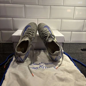 Grå och blå fotbollsskor från Nike - Snygga fotbollsskor från Nike i grått med blå detaljer och orangea inslag på sulan. Skorna har snörning och en strömlinjeformad design för optimal prestanda på planen.