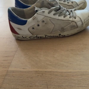 Golden Goose sneakers (never stop dreaming - Snygga Golden Goose sneakers i vitt med en ikonisk stjärndetalj på sidan. Skorna har en cool, sliten look med blå och röda detaljer på hälen. Texten 'Never Stop Dreaming' pryder sulan för en unik touch. Perfekta för en avslappnad stil. 7/10