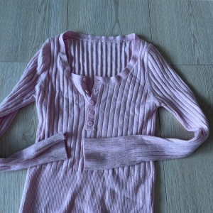 Rosa ribbad tröja med knappar - Säljer en söt rosa ribbad tröja med knappar framtill. Tröjan har långa ärmar och är tillverkad i en mjuk polyesterblandning. Perfekt för en stilren och bekväm look.🩷