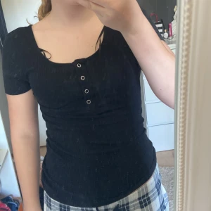 Svart ribbad topp från Pieces - Säljer två svarta ribbad toppar från Pieces i storlek XS. Toppen har korta ärmar och dekorativa knappar framtill. Perfekt för en stilren look. Säljer både i ett köp
