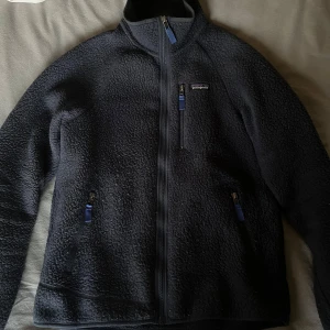 Patagonia fleece - Säljer en svart fleecetröja från Patagonia med dragkedja framtill och tre fickor med dragkedjor. Perfekt för kyliga dagar och outdoor-äventyr. Jackan har en hög krage för extra värme.