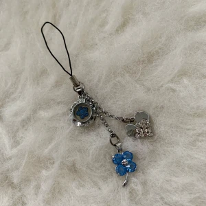 Silverfärgad mobilhänge med blå detaljer - Charmigt mobilhänge i silverfärg med tre hängande berlocker: en blå blomma, ett hjärta med glittriga stenar och en rund berlock med blå blomdetalj. Perfekt för att ge din mobil en personlig touch.