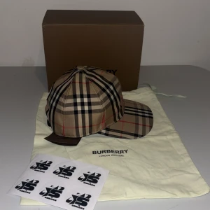 Rutig keps från Burberry - Snygg rutig keps från Burberry i klassiskt beige, svart och rött mönster. Kepsen har justerbar passform och är perfekt för en stilren look. Kommer med originalförpackning och påse. Finns i M L
