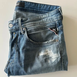 Blå jeans från Replay - Snygga blå jeans från Replay med slitna detaljer. Modellen är Willbi som är som anbass i passformen. Dom är sparsamt använda och i mycket bra skick!