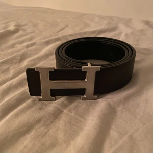 Svart bälte med H-spänne - Snyggt svart bälte med ett stort H-format spänne i silver. Tillverkat i skinn och passar perfekt för att ge en stilren touch till din outfit. Bältet har en klassisk design och är lätt att matcha med olika kläder.