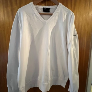Vit tröja från Peak Performance - Säljer en stilren vit tröja från Peak Performance med v-ringning och broderad logga på ärmen. Tröjan har långa ärmar och ribbade muddar vid ärmslut och nederkant. Perfekt för en avslappnad och snygg look!