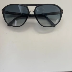 Svarta solglasögon från Tom Ford - Snygga svarta solglasögon från Tom Ford med en klassisk aviatorform. De har mörka linser och en elegant design med gulddetaljer vid gångjärnen. Perfekta för en stilren look.