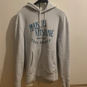 Ljusblå hoodie från Maison Kitsuné - Säljer en stilren vit hoodie från Maison Kitsuné med blå text på framsidan. Den har en klassisk känguruficka och justerbar huva med dragsko. Perfekt för en avslappnad look. Skick 9/10