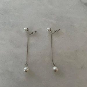 Långa Öronklämma med pärlor - Säljer ett par eleganta långa Öronklämma med vita pärlor i båda ändar. De har en tunn silverfärgad kedja som ger en stilren look. Perfekta för att ge en touch av elegans till din outfit.
