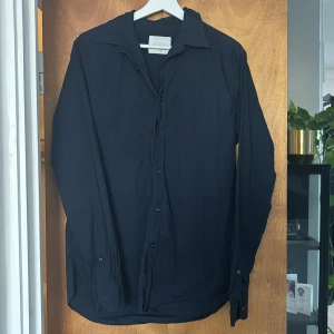 Marinblå skjorta från Oscar Jacobson - Snygg marinblå skjorta från Oscar Jacobson i storlek M. I mycket gott skick. Skjortan är tillverkad i bomull med en stilren design och knappar framtill. Perfekt för en elegant look.