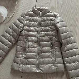 Beige jacka från mango - Snygg beige/grå jacka med horisontella sömmar och hög krage. Jackan har två fickor med dragkedjor och en rak passform. Perfekt för kyliga dagar när du vill ha en stilren look.