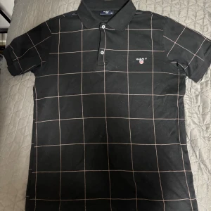 Gant Piké - Svart GANT piké i storlek M med stilrent rutmönster i ljusa linjer. Klassisk krage och två knappar framtill. Broderad GANT-logga med sköld på bröstet. Längd: ca 66 cm, bredd: ca 48 cm.