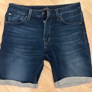 Mörkblå jeansshorts Jack & Jones - Snygga mörkblå jeansshorts från Jack & Jones med klassisk femficksdesign och uppvikta benslut. Shortsen har normal passform, knapp och dragkedja framtill samt detaljerade bakfickor med kontrastsöm. Perfekta för sommarens chill dagar.