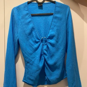 Blå blus  - Snygg blå omlottopp från Gina Tricot med lång ärm och v-ringning. Toppen har en cool ringdetalj framtill med pärlor och lätt rynkad effekt. Materialet är strukturerat och har lite bredare armar nertill. perfekt till jeans eller kjol.