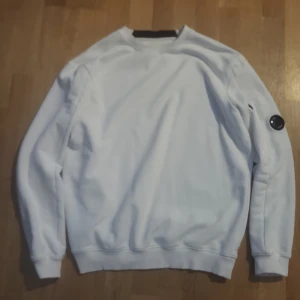 Vit sweatshirt från C.P. Company - Säljer en clean vit sweatshirt från C.P. Company i storlek M. Tröjan har rund hals, ribbade muddar och en ikonisk svart linsdetalj på ärmen. Tillverkad i mjuk bomull som är skön mot huden. Perfekt för dig som gillar stilrena och moderna plagg.