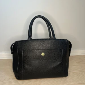 Svart handväska i skinn med guldiga detaljer - Snygg svart handväska från Rizzo i präglat skinn med guldiga metalldetaljer. Väskan har dubbla handtag, dragkedja upptill, ytterfack fram och bak samt ljusbeige foder och flera innerfack. Perfekt storlek för allt du behöver till vardags.