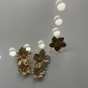 Guldiga blommiga örhängen - Säljer ett par riktigt snygga örhängen i guldton med blommig design. Varje örhänge har tre detaljerade blommor med fina strukturerade blad, perfekt för dig som vill sticka ut med något unikt och trendigt. Passar dig som gillar statement-smycken och blommiga detaljer.
