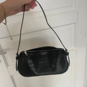 Svart axelväska i croco-imitation - Snygg svart axelväska med croco-mönstrad yta och silvrig dragkedja. Väskan har en stilren, avlång form och ett smalt axelband. Perfekt för dig som gillar trendiga accessoarer med lite edge.