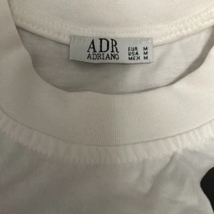 Vit t-shirt med svarta blommor ADR Adriano - Vit t-shirt från ADR Adriano i storlek M med stora svarta blommönster över hela tröjan. Klassisk rund halsringning och korta ärmar. Skön och avslappnad passform, perfekt för dig som gillar en unik och trendig look. Pris kan diskuteras.