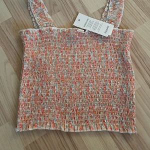 Blommig linnetopp från Kappahl - Supersöt linnetopp från Kappahl med blommigt mönster i orange, rosa och vitt. Toppen har smockad struktur, volangkant upptill och nertill samt breda axelband. Perfekt för varma dagar och ger en fräsch sommarkänsla.