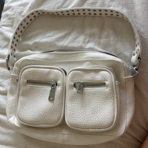 Vit noella skinn väska - Säljer en vit axelväska i skinn med två framfickor med dragkedja och en bred, justerbar axelrem med coola prickdetaljer. Väskan har silvriga metalldetaljer och en avtagbar rem för extra flexibilitet. Perfekt för dig som vill ha både stil och funktion.