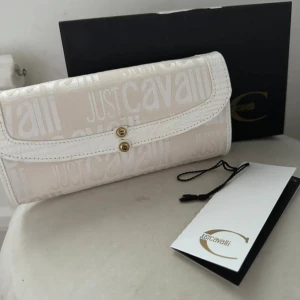 Beige/créamvit plånbok från Just Cavalli - Snygg beige plånbok från Just Cavalli med logomönster över hela ytan. Plånboken har vita detaljer i med struktur, guldiga knappar och flera fack inuti. Perfekt för dig som vill ha en stilren och lyxig accessoar