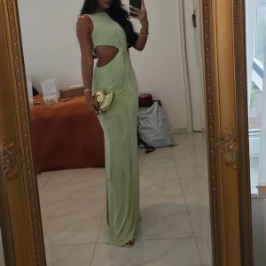 Ljusgrön långklänning med cut-outs - Säljer en ljusgrön långklänning med snygga cut-outs i midjan och ett knyt framtill. Klänningen är ärmlös och har en slim passform som framhäver figuren. Perfekt för dig som vill sticka ut med en modern och fräsch look.