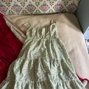 Grön blommig långklänning med smala axelband - Säljer en söt grön långklänning med vitt blommönster och smala axelband. Klänningen har volanger och en luftig passform, perfekt för varma dagar. Materialet känns lätt och bekvämt mot huden. Passar dig som gillar romantisk och chill stil.