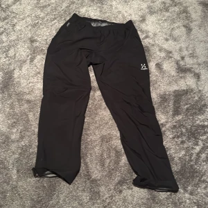 Svarta täckbyxor från Haglöfs - Svarta täckbyxor från Haglöfs med elastisk midja och meshfoder på insidan. Byxorna är raka i modellen och tillverkade i ett lätt syntetmaterial som passar perfekt för utomhusaktiviteter. Diskret Haglöfs-logga på låret.