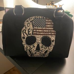 Svart väska - Unik svart väska med dödskalle motiv och lite svarta nitar. Finns ett längre band till väskan. Köpt i USA och nästan oanvänd 