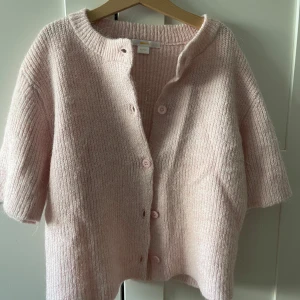 Ljusrosa stickad kofta från Gina Tricot - Supermysig ljusrosa stickad kofta från Gina Tricot med korta ärmar och rund hals. Koftan har ribbad struktur och knäpps med matchande rosa knappar framtill. Perfekt för lager-på-lager och chill dagar.