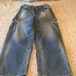 Diesel Widee-J-SP1 wide jeans blå - Blå jeans från Diesel, modell Widee-J-SP1, med bred passform och elastisk midja. Jeansen har slitna detaljer och hål vid knäna för en cool, avslappnad look. Klassisk femficksdesign och dragkedja. Perfekta för dig som gillar loose och wide fit. Längd from midjan ner: 96,5 cm. Midjan: 37 cm