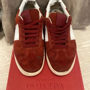 Valentino Garavani röda sneakers mocka - Snygga sneakers från Valentino Garavani i röd mocka med vita skinnpaneler och vita detaljer på hälen. Klassisk låg modell med snörning och cool sula med kamouflagemönster undertill. Perfekta för dig som vill sticka ut med exklusiva och trendiga skor. Skon är ungefär 23 cm inuti vilket motsvara storlek 36-37.