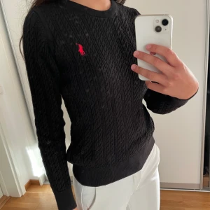 Svart kabelstickad tröja från Polo Ralph Lauren - Svart kabelstickad tröja från Polo Ralph Lauren med klassisk röd logga broderad på bröstet. Rund halsringning och långa ärmar. Mjuk och skön som passar perfekt till jeans. Snygg ribbad kant vid ärmslut och nederkant.