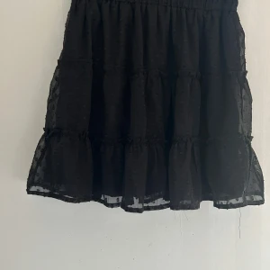 Svart prickig volangkjol - Supersöt svart kortkjol med volanger och prickigt mönster i mesh. Kjolen har en luftig och lätt känsla med flera lager och resår i midjan. Perfekt för dig som gillar att sticka ut med detaljer och älskar volanger.