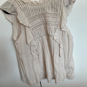 Beige blus med volanger från Holly & Whyte - Supersöt beige blus från Holly & Whyte med volanger längs axlarna och framstycket. Blusen har broderade spetsdetaljer och tunna vertikala sömmar upptill. Luftig och lätt i bomull, perfekt för ljusa dagar.