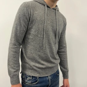 Grå stickad hoodie med huva - Snygg grå hoodie med huva och snörning, tillverkad i mjukt stickat MERINOULL & KASHMIR. Klassisk passform som funkar till jeans eller andra favoritplagg. Perfekt för dig som gillar enkel och stilren look.