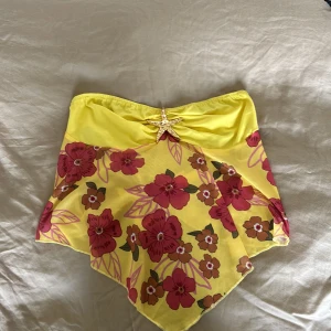 Gul blommig bandeau tubtopp - Superfin gul bandeau tubtopp med råsa blommor och bladmönster. Toppen är asymmetrisk och har en dekorativ sjöstjärna 