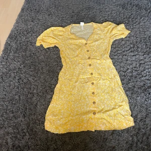 Gul blommig kortklänning från H&M - Säljer en söt gul kortklänning från H&M med vita blommor, v-ringad hals och knappar framtill. Klänningen har korta puffärmar med resår och är gjord i ett lätt och skönt material, perfekt för soliga dagar. Passformen är normal och den har en feminin siluett.