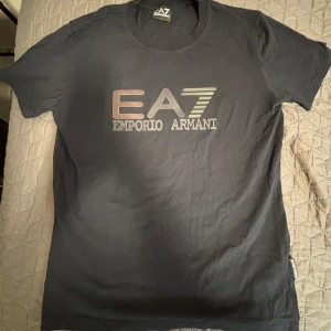 Emporio Armani T-shirt - Svart EA7 Emporio Armani T-shirt med ett tryck på bröstet som är värmereaktivt och byter färg på värme. Klassisk rund hals och korta ärmar. Mjuk och skön bomullskvalitet, perfekt för en clean och stilren look. Snygg logotypdetalj på sidan.