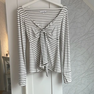Randig långärmad topp Design by Si - Supersnygg långärmad topp från Design by Si i vitt med svarta ränder. Djup v-ringning och knyt framtill ger en trendig look. Toppens material är mjukt. Har ändast används 1 gång