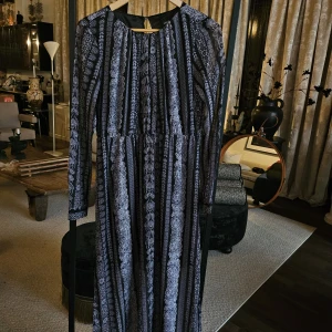 Svart långklänning med vitt mönster - Långklänning från Monki i svart med vitt, detaljerat mönster i vertikala ränder. Klänningen har lång ärm, rund halsringning och ett flowy fall. Perfekt för dig som gillar bohemiska vibes och vill sticka ut med ett unikt print.