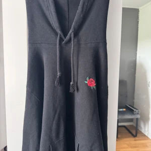 Svart hoodie med rosbroderi - Svart hoodie med huva och snörning framtill. På bröstet finns ett broderat motiv av en röd ros med gröna blad. Tröjan har en klassisk känguruficka och är gjord i mjuk bomullsmix. Perfekt för en chill och stilren look.