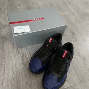 Blå och svarta sneakers från Prada - Snygga Prada Cups i blått och svart med glansiga paneler och meshdetaljer. Skorna har svart snörning, rund tå och en chunky svart sula. Röd Prada-logga på plösen ger en exklusiv touch. Perfekta för dig som gillar statement-sneakers.