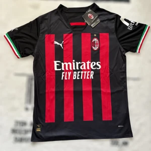 AC Milan Puma fotbollströja röd/svart - AC Milan matchtröja från Puma med klassiska röda och svarta ränder, klubbmärke på bröstet och sponsortryck. Tröjan har korta ärmar med italienska flaggans färger på muddarna och är tillverkad i lätt och ventilerande Drycell-material.